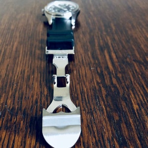 Puede incluir: Primer plano de una correa de reloj plateada con cierre. La correa es de metal y tiene un acabado pulido.