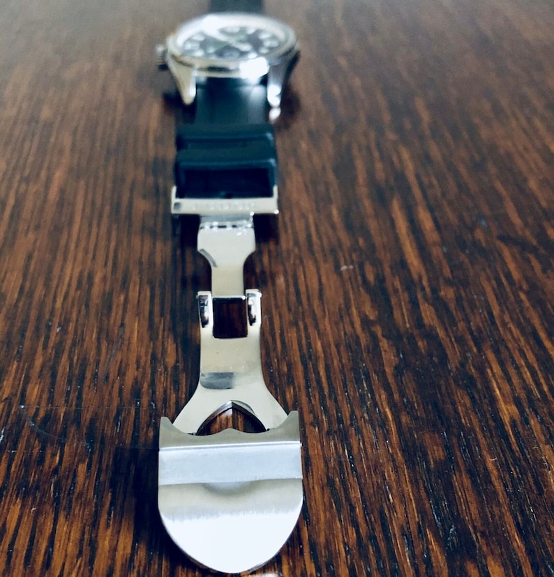 Puede incluir: Primer plano de una correa de reloj plateada con cierre. La correa es de metal y tiene un acabado pulido.