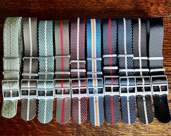 May include: A collection of nylon watch straps in various colors and patterns. The straps feature stripes in red, white, blue, and orange, as well as solid colors like olive green and black. Each strap has a silver-colored buckle.