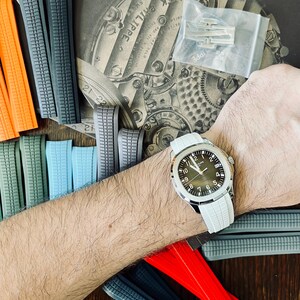 May include: A wristwatch with a white strap and a dark face is shown on a person's wrist. Various coloured watch straps in orange, green, blue, grey, and red are arranged around the watch. A package of watch clasps is also visible.