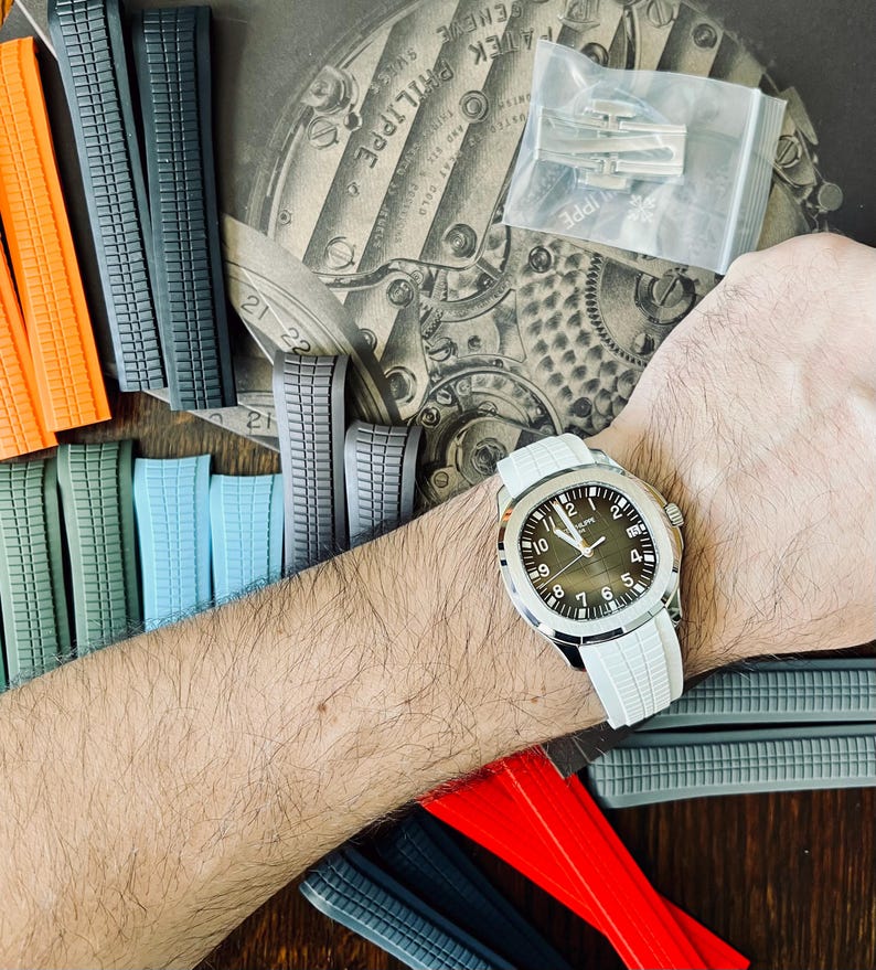 May include: A wristwatch with a white strap and a dark face is shown on a person's wrist. Various coloured watch straps in orange, green, blue, grey, and red are arranged around the watch. A package of watch clasps is also visible.