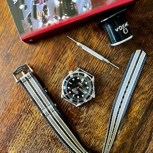 Puede incluir: Un reloj de pulsera con esfera negra y caja plateada, junto a una correa a rayas. El reloj está junto a una herramienta de reparación de relojes, una lupa y un libro con el logotipo de Omega. La correa es negra, gris y beige.