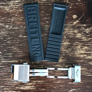 May include: A black rubber watch strap with the word "BREITLING" embossed on it. The strap is attached to a silver metal watch buckle with the word "BREITLING" and the year "1884" engraved on it.