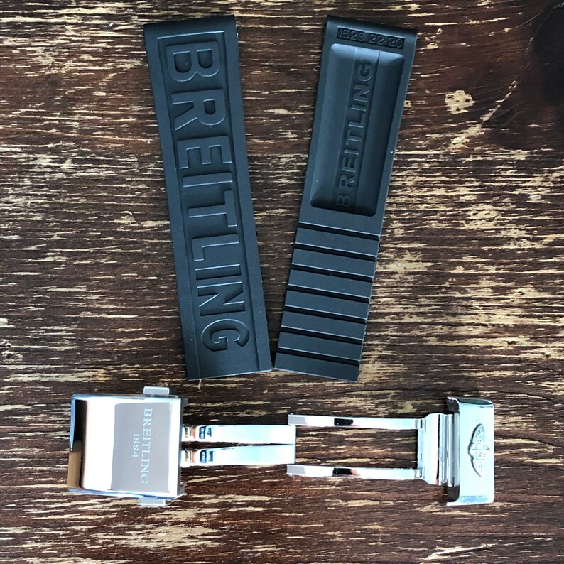 May include: A black rubber watch strap with the word "BREITLING" embossed on it. The strap is attached to a silver metal watch buckle with the word "BREITLING" and the year "1884" engraved on it.