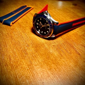 Puede incluir: Un reloj de pulsera de acero inoxidable con esfera negra, bisel azul y correa roja y azul. El reloj tiene agujas e indicadores dorados. Se ve una correa de reloj azul y naranja adicional.