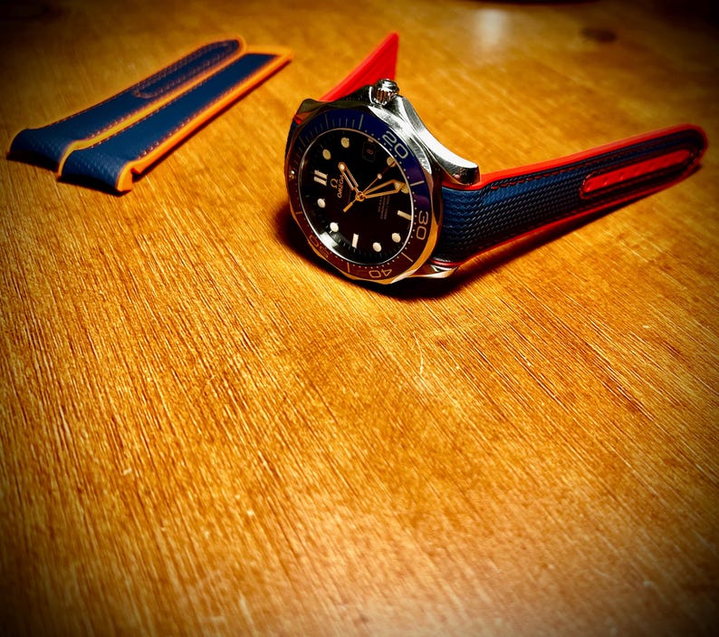 Puede incluir: Un reloj de pulsera de acero inoxidable con esfera negra, bisel azul y correa roja y azul. El reloj tiene agujas e indicadores dorados. Se ve una correa de reloj azul y naranja adicional.