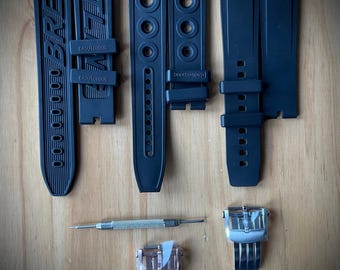 May include: Black watch straps and clasps with a small screwdriver. The straps have the word "BREITLING" and "GAUTCHOUX" printed on them. The clasps are silver and rose gold.