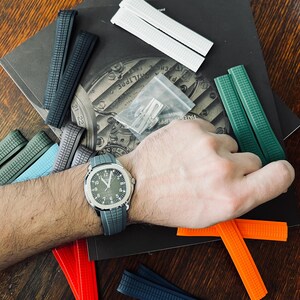 May include: A wristwatch with a grey strap and a green face is shown with a selection of coloured watch straps. The straps are in shades of green, blue, orange, white, and black. A small bag of watch strap pins is also visible.