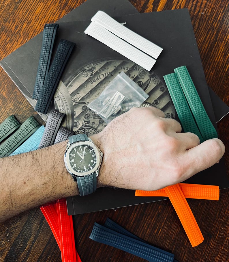 May include: A wristwatch with a grey strap and a green face is shown with a selection of coloured watch straps. The straps are in shades of green, blue, orange, white, and black. A small bag of watch strap pins is also visible.