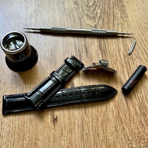 May include: A black leather watch strap with a crocodile pattern, a silver watch buckle, a small magnifying glass with the word "OMEGA" printed on it, and a small black tube with the word "OMEGA" printed on it.