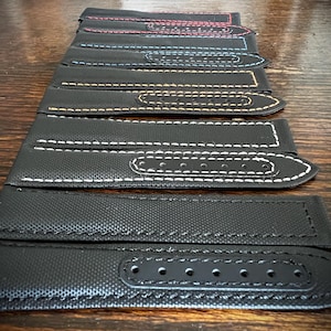 Puede incluir: Una colección de correas de reloj negras con costuras en contraste en varios colores, incluyendo rojo, azul, amarillo y blanco. Las correas están hechas de un material texturizado y tienen múltiples agujeros para ajustar.
