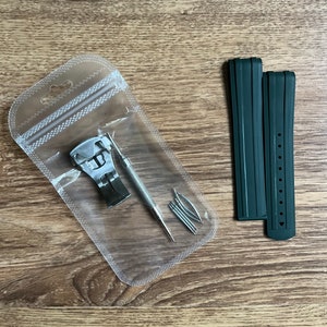 Puede incluir: Un juego de dos correas de reloj verde oscuro con una herramienta de banda de reloj plateada y pasadores de resorte en una bolsa de pl&aacute;stico transparente.