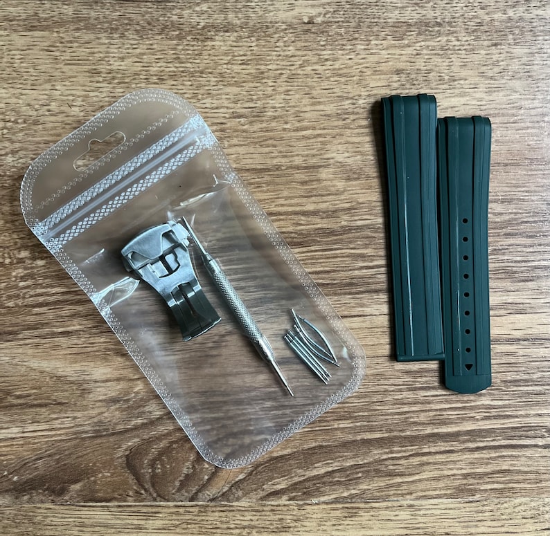 Puede incluir: Un juego de dos correas de reloj verde oscuro con una herramienta de banda de reloj plateada y pasadores de resorte en una bolsa de pl&aacute;stico transparente.