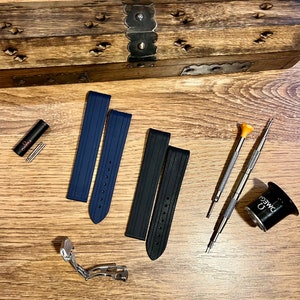 Puede incluir: Dos correas de reloj, una azul y otra negra, con un cierre de banda de reloj plateado. Las correas est&aacute;n hechas de un material de goma texturizado. Tambi&eacute;n hay dos peque&ntilde;os destornilladores, una lupa con la palabra "OMEGA" impresa en ella y una peque&ntilde;a herramienta roja con la palabra "OMEGA" impresa en ella.