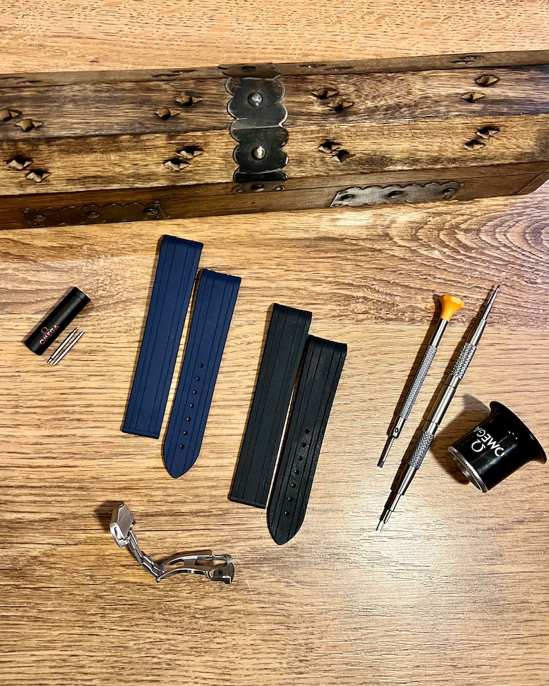 Puede incluir: Dos correas de reloj, una azul y otra negra, con un cierre de banda de reloj plateado. Las correas est&aacute;n hechas de un material de goma texturizado. Tambi&eacute;n hay dos peque&ntilde;os destornilladores, una lupa con la palabra "OMEGA" impresa en ella y una peque&ntilde;a herramienta roja con la palabra "OMEGA" impresa en ella.