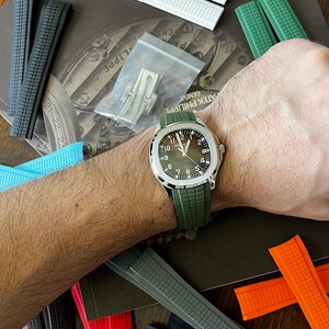 May include: A wristwatch with a green strap is shown, alongside a selection of watch straps in colours including white, black, grey, blue, and orange. The watch features a silver case and a dark green face. A small bag of watch components is also visible.