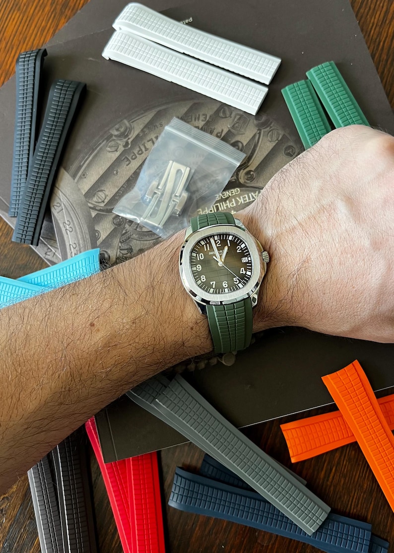 May include: A wristwatch with a green strap is shown, alongside a selection of watch straps in colours including white, black, grey, blue, and orange. The watch features a silver case and a dark green face. A small bag of watch components is also visible.