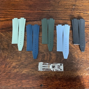 May include: An assortment of watch straps in various colors, including light blue, navy, olive green, and gray. The straps are made of a textured material and are arranged on a wooden surface. A silver watch clasp is also present.