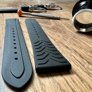 Puede incluir: Dos correas de reloj negras con diferentes texturas. Una correa tiene una superficie lisa con agujeros, mientras que la otra tiene una superficie texturizada con un patr&oacute;n de l&iacute;neas diagonales. Las correas est&aacute;n sobre una superficie de madera.