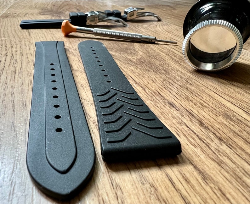 Puede incluir: Dos correas de reloj negras con diferentes texturas. Una correa tiene una superficie lisa con agujeros, mientras que la otra tiene una superficie texturizada con un patr&oacute;n de l&iacute;neas diagonales. Las correas est&aacute;n sobre una superficie de madera.