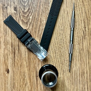 Puede incluir: Dos correas de reloj negras con un patr&oacute;n texturizado y una herramienta plateada para reparaciones de relojes sobre una superficie de madera.
