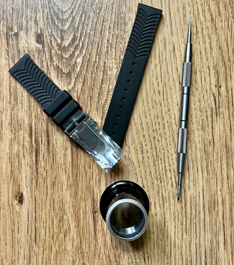 Puede incluir: Dos correas de reloj negras con un patr&oacute;n texturizado y una herramienta plateada para reparaciones de relojes sobre una superficie de madera.