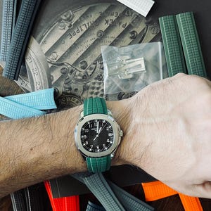 May include: A wristwatch with a black face and silver casing is worn on a wrist with a green watch strap. Various coloured watch straps in grey, blue, white, and red are scattered around the watch. The watch face displays the numbers 1 through 12.