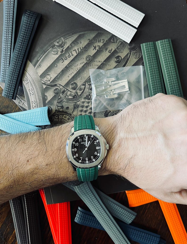 May include: A wristwatch with a black face and silver casing is worn on a wrist with a green watch strap. Various coloured watch straps in grey, blue, white, and red are scattered around the watch. The watch face displays the numbers 1 through 12.