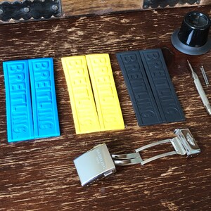 May include: A set of watch straps with the word "Breitling" printed on them. The straps are in blue, yellow, and black. There is also a silver watch buckle with the word "Breitling" engraved on it.