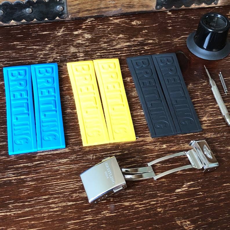 May include: A set of watch straps with the word "Breitling" printed on them. The straps are in blue, yellow, and black. There is also a silver watch buckle with the word "Breitling" engraved on it.