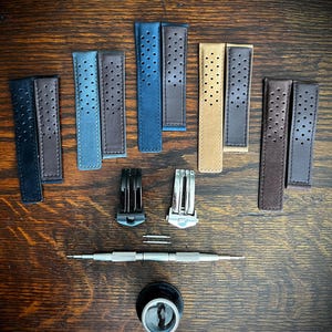 Può includere: Un assortimento di cinturini per orologi in pelle di vari colori, tra cui nero, blu e marrone, con design perforati. Sono inclusi anche chiusure per orologi, barrette a molla, uno strumento per la riparazione di orologi e una lente d'ingrandimento, disposti su una superficie di legno.