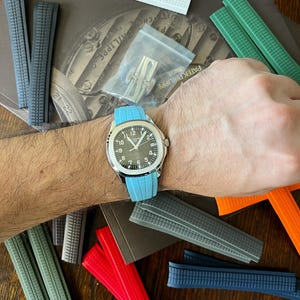 May include: A wristwatch with a silver case and a black dial, featuring white hour markers and hands, is displayed with a light blue band. Various coloured watch straps in white, grey, green, red, and blue are scattered around the watch.