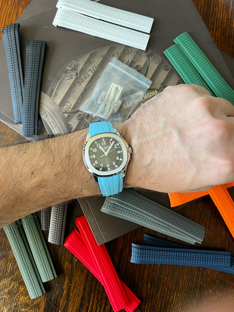 May include: A wristwatch with a silver case and a black dial, featuring white hour markers and hands, is displayed with a light blue band. Various coloured watch straps in white, grey, green, red, and blue are scattered around the watch.