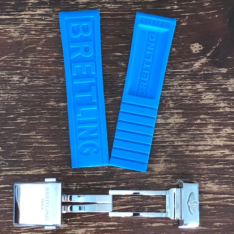May include: Two blue rubber watch straps with the word "BREITLING" printed on them. The straps are for a watch with a 22mm lug width. A silver watch buckle with the word "BREITLING 1884" engraved on it.