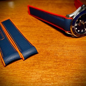 Puede incluir: Un reloj de lujo con esfera azul y bisel plateado, junto con una correa azul y naranja. La correa es de material texturizado con costuras naranjas. El reloj y la correa est&aacute;n sobre una superficie de madera.