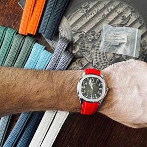 May include: A silver wristwatch with a black face and white numerals is worn on a wrist with a red band. Various coloured watch straps in orange, grey, blue, and white are displayed. The watch face reads "PATEK PHILIPPE".