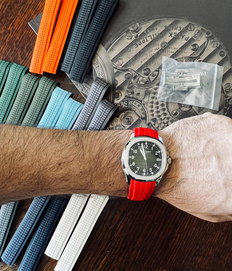 May include: A silver wristwatch with a black face and white numerals is worn on a wrist with a red band. Various coloured watch straps in orange, grey, blue, and white are displayed. The watch face reads "PATEK PHILIPPE".