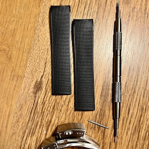 Puede incluir: Dos correas de reloj negras con una superficie texturizada, un cierre de banda de reloj plateado y una herramienta de banda de reloj plateada.