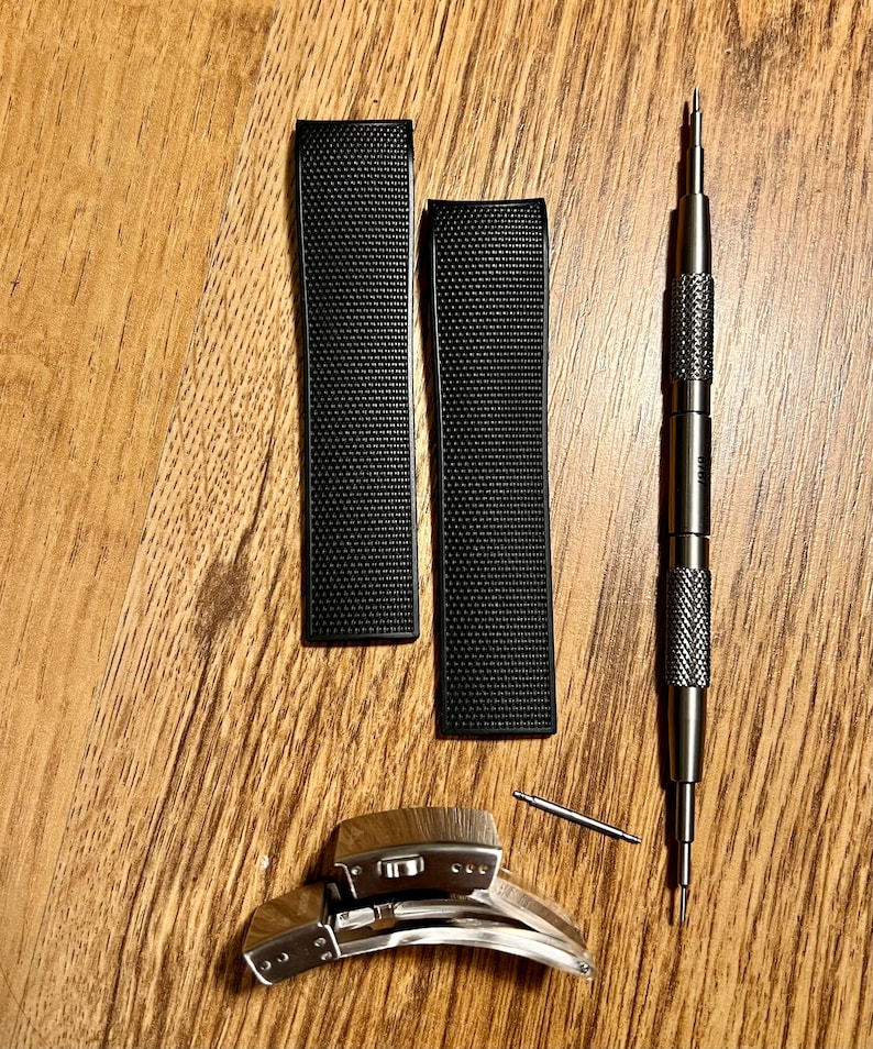 Puede incluir: Dos correas de reloj negras con una superficie texturizada, un cierre de banda de reloj plateado y una herramienta de banda de reloj plateada.