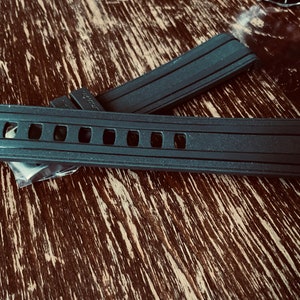 May include: Two black rubber watch bands with a textured surface and multiple holes. The band on top has a small logo on the side.
