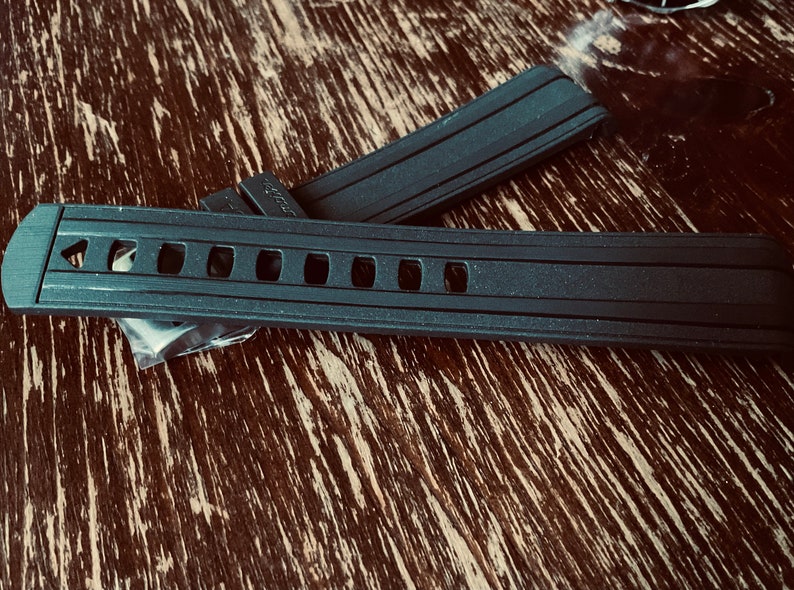 May include: Two black rubber watch bands with a textured surface and multiple holes. The band on top has a small logo on the side.