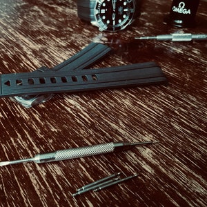 May include: A black watch strap with a buckle and a row of holes. The strap is lying on a wooden surface with a small screwdriver and spring bars.