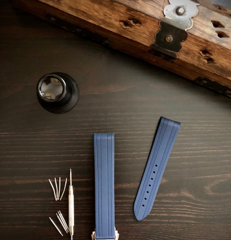 Puede incluir: Dos correas de reloj azules con una hebilla plateada. Las correas est&aacute;n hechas de un material de goma texturizado y tienen un extremo ligeramente puntiagudo. Tambi&eacute;n hay varias herramientas peque&ntilde;as de plata sobre una superficie de madera oscura.