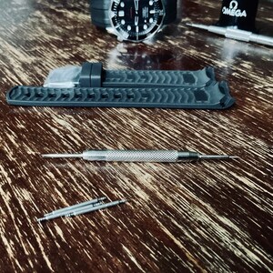 May include: A close-up of a black watch strap with a textured surface. The strap is lying on a wooden surface with a silver watch tool and four silver spring bars.