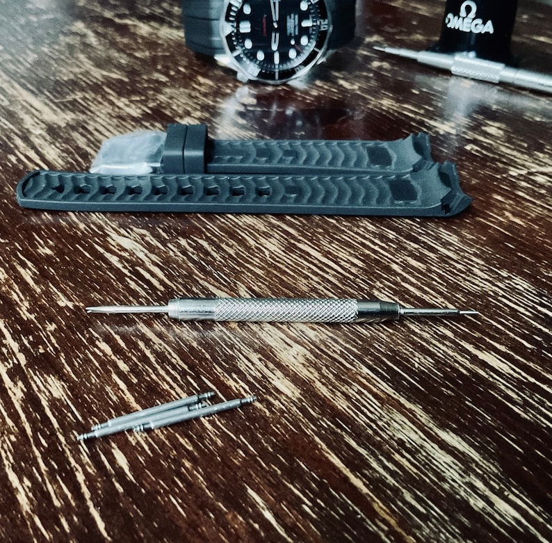 May include: A close-up of a black watch strap with a textured surface. The strap is lying on a wooden surface with a silver watch tool and four silver spring bars.