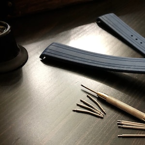 Puede incluir: Una correa de reloj de silicona azul con hebilla plateada y una herramienta para quitar los pasadores del reloj. La herramienta est&aacute; sobre una superficie de madera oscura con varios pasadores de reloj esparcidos alrededor.