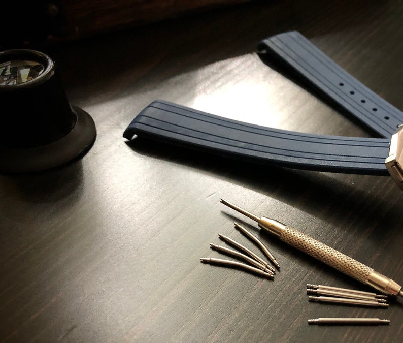 Puede incluir: Una correa de reloj de silicona azul con hebilla plateada y una herramienta para quitar los pasadores del reloj. La herramienta est&aacute; sobre una superficie de madera oscura con varios pasadores de reloj esparcidos alrededor.