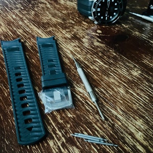 May include: Two black watch bands with a silver buckle, a silver watch tool, and four silver watch pins on a wooden surface.