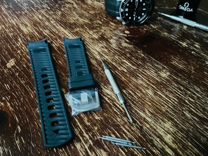May include: Two black watch bands with a silver buckle, a silver watch tool, and four silver watch pins on a wooden surface.