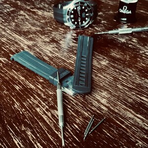 May include: A close-up of a black watch band with a silver watch face and silver watch hands. The watch band is laying on a wooden surface with a silver watch tool and a small bottle of oil labeled "OMEGA".